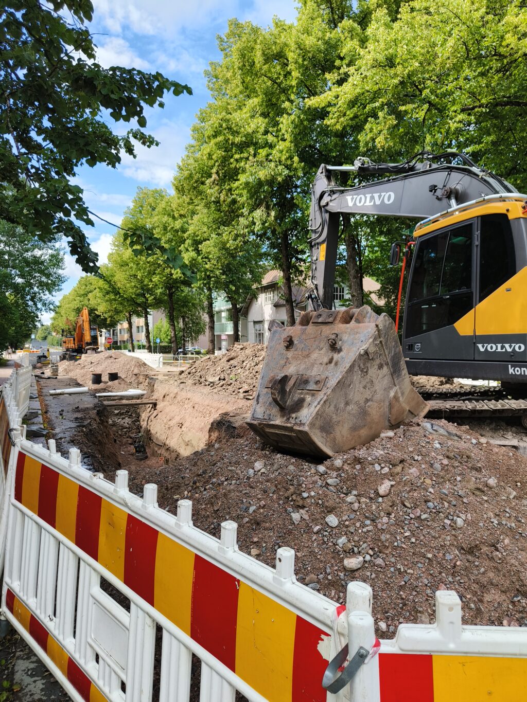 Työmaa-aitojen sisällä kuoppa, jonka kaivuri on kaivanut. Kuopassa vesiputkia ja viemäri.